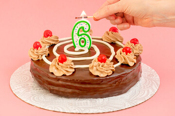 Birthday chocolate cake with burning candle in the form of number Six. Homemade cake decorated with chocolate cream and cherries close up on pink background