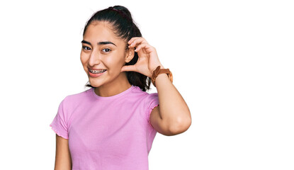 Fototapeta premium Hispanic teenager girl with dental braces wearing casual clothes smiling with hand over ear listening an hearing to rumor or gossip. deafness concept.