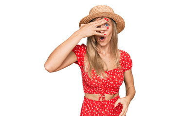 Beautiful caucasian woman wearing summer hat peeking in shock covering face and eyes with hand,...