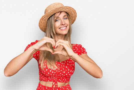 Beautiful caucasian woman wearing summer hat smiling in love doing heart symbol shape with hands. romantic concept.