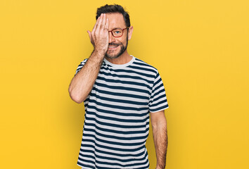 Middle age man wearing casual clothes and glasses covering one eye with hand, confident smile on face and surprise emotion.