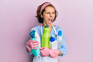 Young brunette woman wearing cleaner apron holding cleaning products angry and mad screaming...