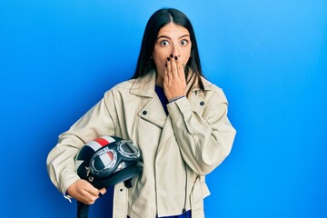 Young hispanic woman holding motorcycle helmet covering mouth with hand, shocked and afraid for mistake. surprised expression