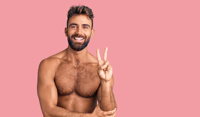 Young hispanic man wearing swimwear shirtless smiling with happy face winking at the camera doing victory sign. number two.