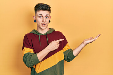 Young caucasian boy with ears dilation wearing casual sweatshirt amazed and smiling to the camera while presenting with hand and pointing with finger.