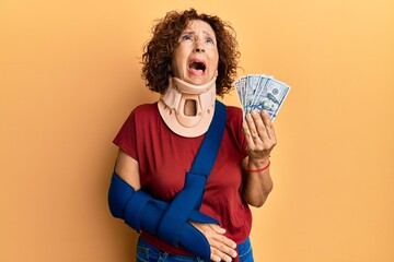 Beautiful middle age mature woman wearing cervical collar and sling holding insurance dollars angry...