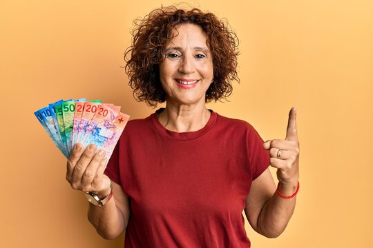 Beautiful middle age mature woman holding swiss franc banknotes smiling with an idea or question pointing finger with happy face, number one