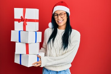 Beautiful hispanic woman wearing christmas hat and holding gifts winking looking at the camera with...