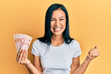 Beautiful hispanic woman holding 10 singapore dollars banknotes screaming proud, celebrating victory and success very excited with raised arm