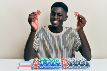 Young african american man playing poker holding casino chips winking looking at the camera with...