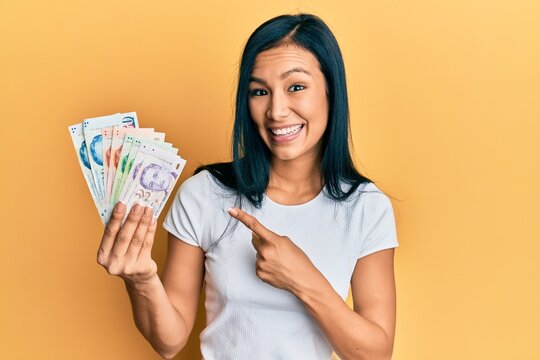 Beautiful hispanic woman holding singapore dollars banknotes smiling happy pointing with hand and finger