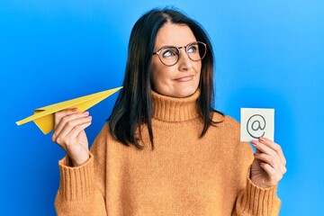 Middle age brunette woman holding email symbol and paper plane smiling looking to the side and...