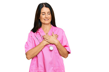 Middle age brunette woman wearing doctor uniform and stethoscope smiling with hands on chest with closed eyes and grateful gesture on face. health concept.