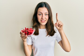 Young brunette woman holding red peppers smiling with an idea or question pointing finger with happy face, number one