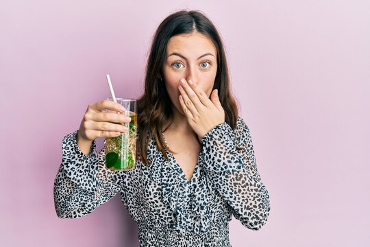 Young brunette woman drinking mojito glass covering mouth with hand, shocked and afraid for mistake. surprised expression