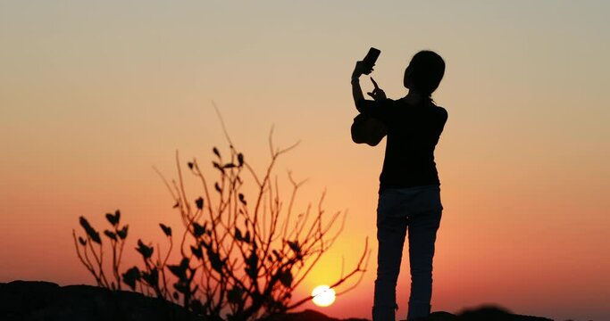 silhouette young woman taking pictures of sunset with her smartphone