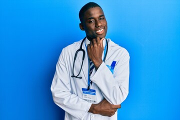 Young african american man wearing doctor uniform looking confident at the camera with smile with crossed arms and hand raised on chin. thinking positive.