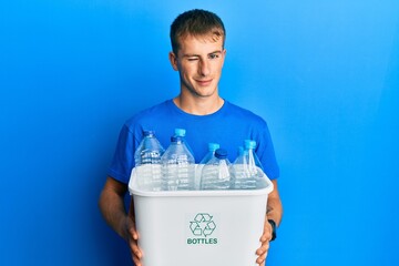 Young caucasian man holding recycling wastebasket with plastic bottles winking looking at the...