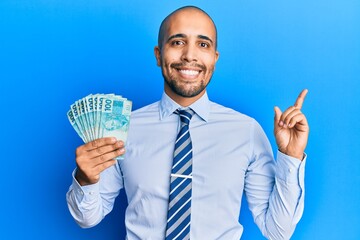 Hispanic adult man holding 100 brazilian real banknotes smiling happy pointing with hand and finger to the side