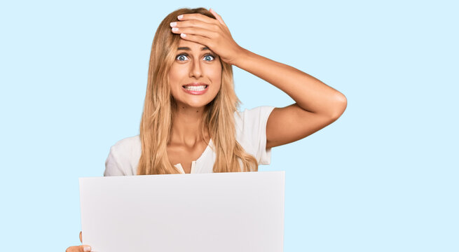 Beautiful blonde young woman holding blank empty banner stressed and frustrated with hand on head, surprised and angry face