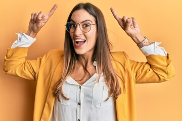 Young beautiful woman wearing business style and glasses smiling amazed and surprised and pointing up with fingers and raised arms.