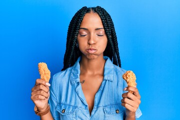 Beautiful hispanic woman eating chicken wings puffing cheeks with funny face. mouth inflated with...