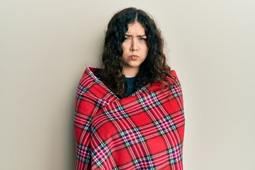 Young brunette woman with curly hair wrapped in a red warm red blanket skeptic and nervous,...