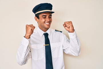 Young hispanic man wearing airplane pilot uniform very happy and excited doing winner gesture with arms raised, smiling and screaming for success. celebration concept.