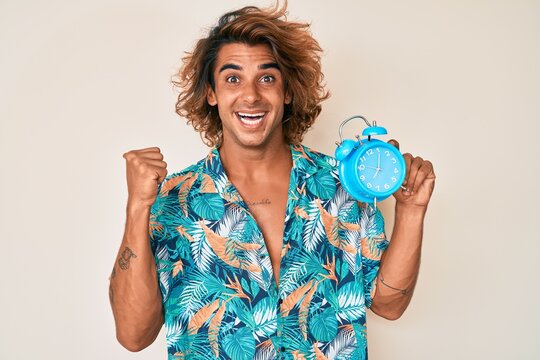 Young hispanic man wearing summer style holding alarm clock pointing thumb up to the side smiling happy with open mouth