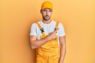 Young handsome man wearing handyman uniform over yellow background pointing with hand finger to the side showing advertisement, serious and calm face