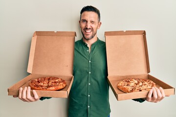 Handsome man with beard holding two italian pizzas winking looking at the camera with sexy...