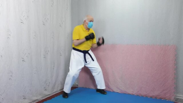 Old man athlete with black pads on his arms and wearing a medical mask trains dodges and punches