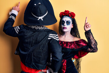 Young couple wearing mexican day of the dead costume over yellow posing backwards pointing ahead with finger hand