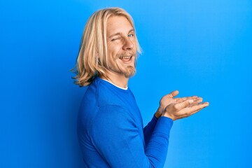 Caucasian young man with long hair presenting with open palms, holding something winking looking at...