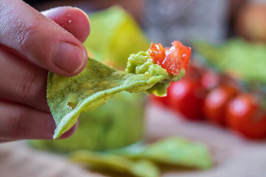 Selective Focus On Hand Holding Green, Avocado Flavored, Tortilla Chip With Fresh Homemade Guacamole Dip. Gluten Free Traditional Mexican Cuisine Or Dish. Cherry Tomatoes On Top And In Background.