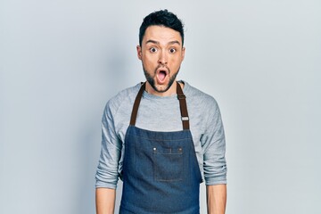 Young hispanic man wearing apron and gloves scared and amazed with open mouth for surprise, disbelief face
