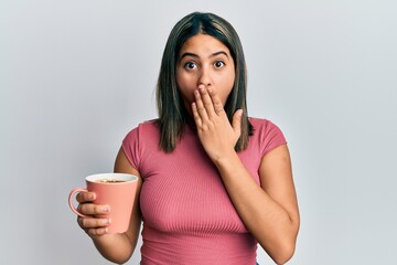 Young latin woman holding coffee covering mouth with hand, shocked and afraid for mistake....