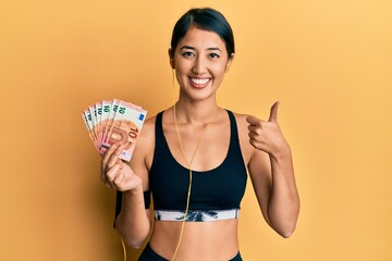 Beautiful asian young sport woman holding bunch of 50 euro banknotes smiling happy and positive,...