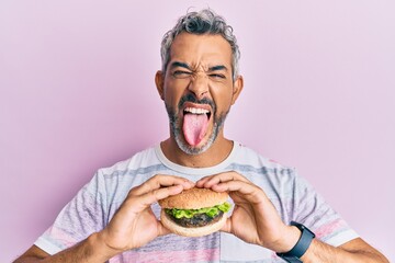 Middle age grey-haired man eating a tasty classic burger sticking tongue out happy with funny expression.