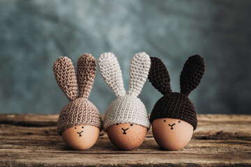 Three Easter eggs in crochet hats with bunny ears on old wooden table - obrazy, fototapety, plakaty