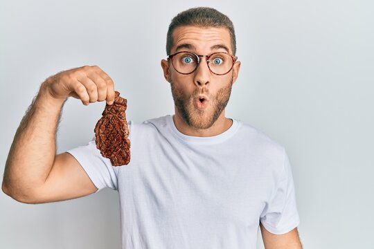 Young caucasian man holding beef steak scared and amazed with open mouth for surprise, disbelief face