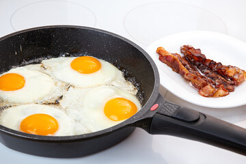 A nutritious breakfast of fried eggs and crispy bacon. The process of cooking a group of eggs in a pan. Crispy bacon on a plate. Idea for a quick, hearty American or British breakfast.