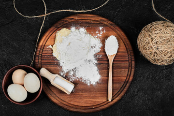 Flour and eggs on wooden board with spoons on black background
