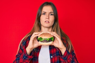 Beautiful caucasian woman eating a tasty classic burger clueless and confused expression. doubt...