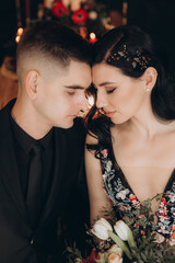 Wedding of a beautiful couple in a black dress, a bouquet of marsala color, at a festive table with candles and a cake against a black wall