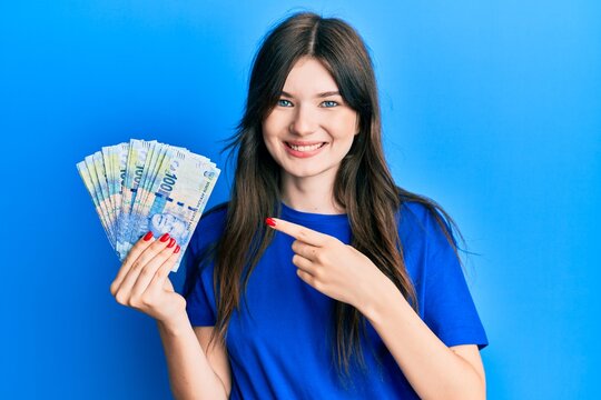 Young beautiful caucasian girl holding south african 100 rand banknotes smiling happy pointing with hand and finger