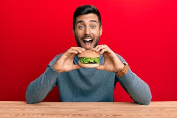 Handsome hispanic man eating a tasty classic burger smiling and laughing hard out loud because funny crazy joke.