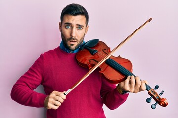 Young hispanic musician man playing violin in shock face, looking skeptical and sarcastic,...
