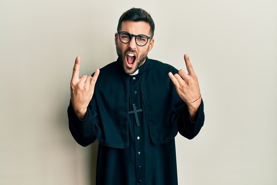 Young hispanic man wearing priest uniform standing over white background shouting with crazy expression doing rock symbol with hands up. music star. heavy concept.