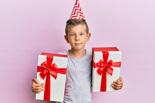 Adorable caucasian kid wearing birthday hat holding presents relaxed with serious expression on face. simple and natural looking at the camera.